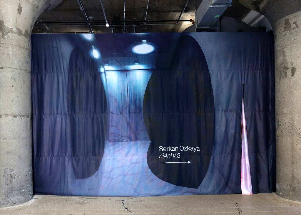 A heavy curtain hangs between two large cylindrical cement pillars. On the curtain is printed an image of the artwork inside with the artist's name and the title of the artwork. A slit in the fabric is open on the right side