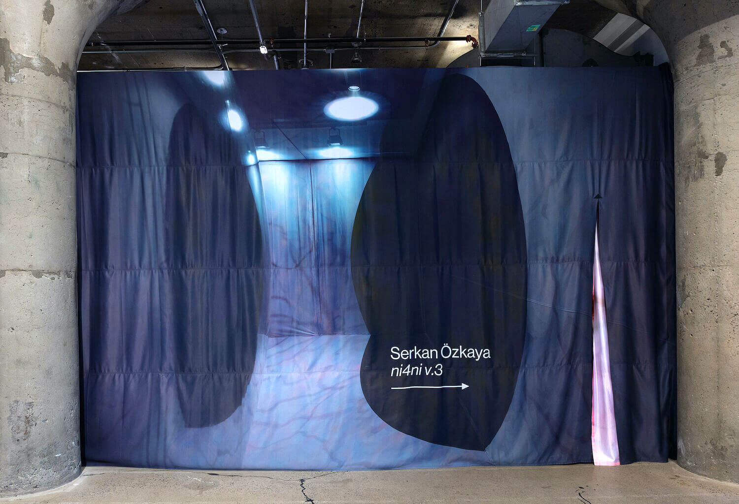 A heavy curtain hangs between two large cylindrical cement pillars. On the curtain is printed an image of the artwork inside with the artist's name and the title of the artwork. A slit in the fabric is open on the right side